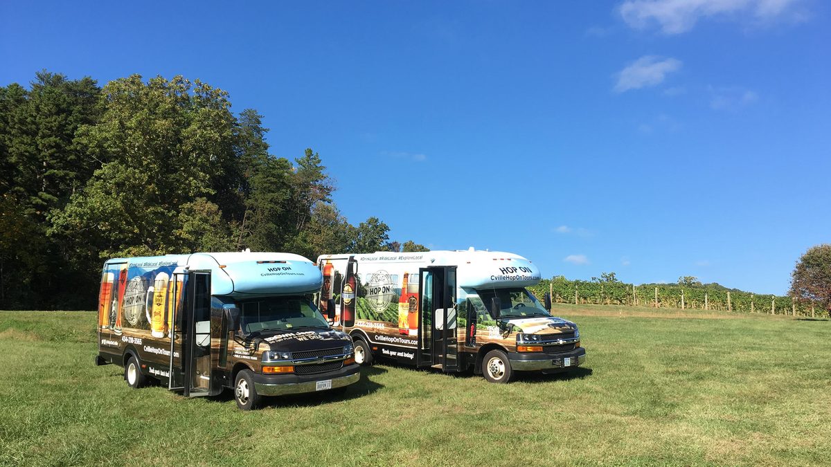 Two Hop On shuttles in their photo wraps, doors open, parked in a mown field beside vineyard rows under a clear blue sky